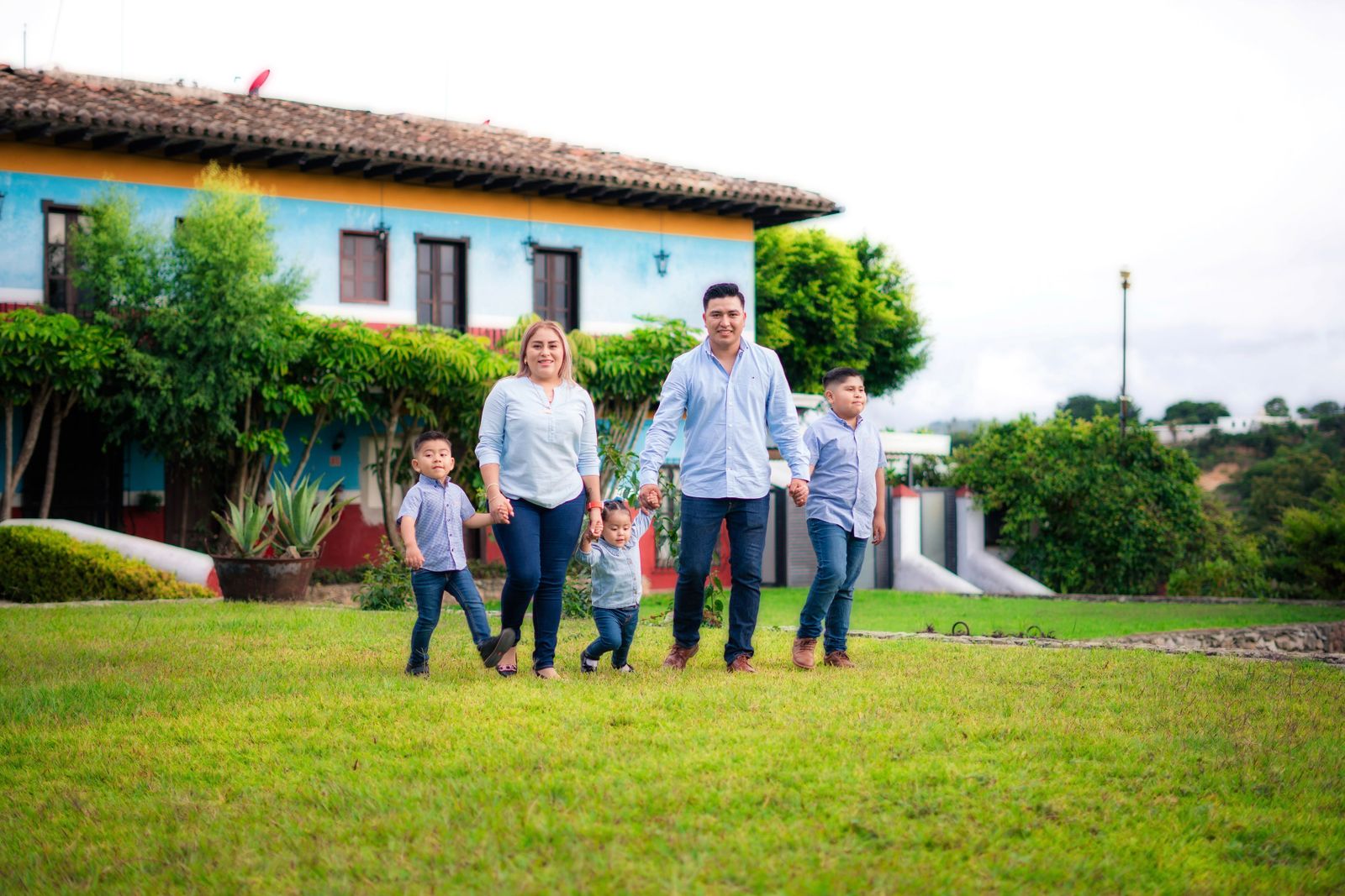 A family of five (mother, father, three young boys) holding hands and walking together across a green lawn in front of a colorful single-story home with a tiled roof and landscaped garden; bright…