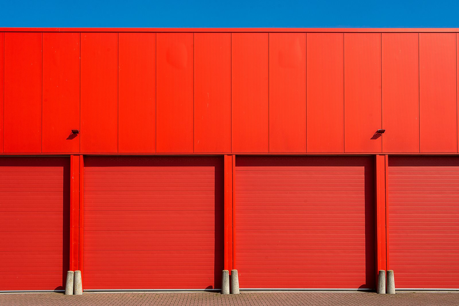Row of four vivid red commercial overhead sectional doors on a red-paneled industrial building with concrete guard bollards, against a deep blue sky.